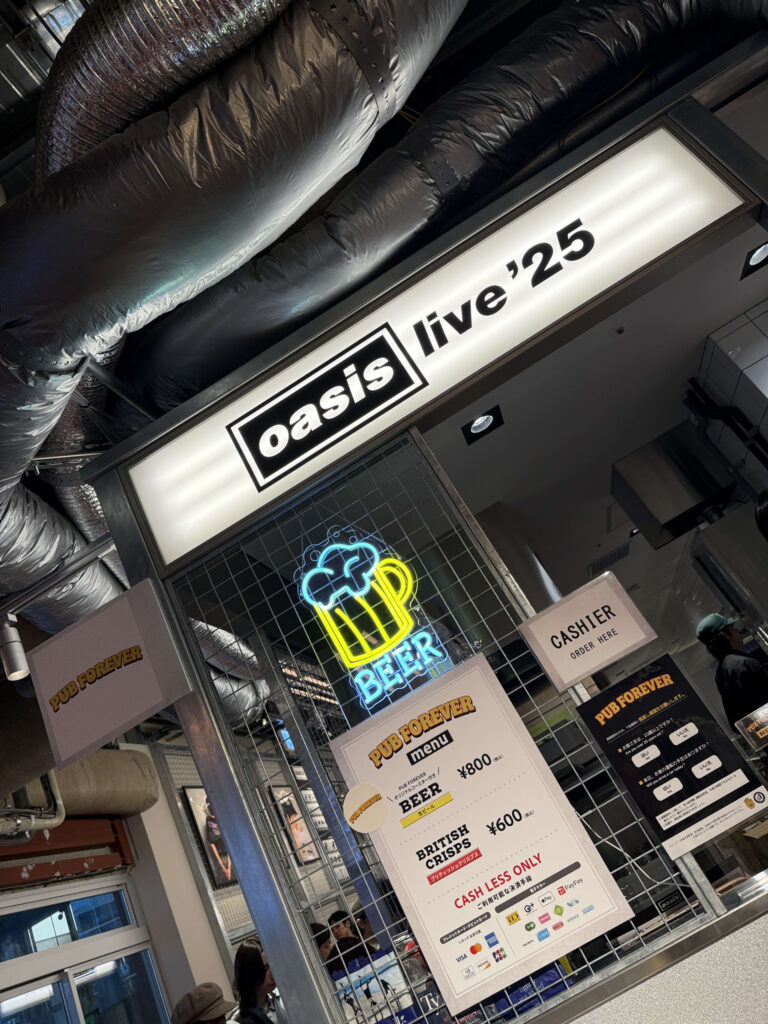 「oasis live '25」イベント会場のパブ「PUB FOREVER」の入り口。むき出しのダクトが天井を走り、ライトアップされた「oasis」の看板とビールのネオンサインがインダストリアルな雰囲気を醸し出している。