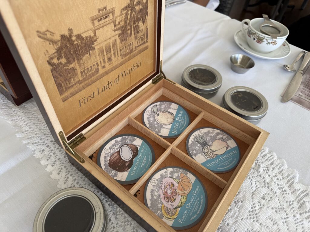 An open wooden tea box on a white lace tablecloth, displaying four tins of Hawaiian tea: 'Sweet Peach,' 'Mango Pineapple,' 'Hawaiian Coconut,' and 'Passion Orange Guava.' A teacup is visible in the background.