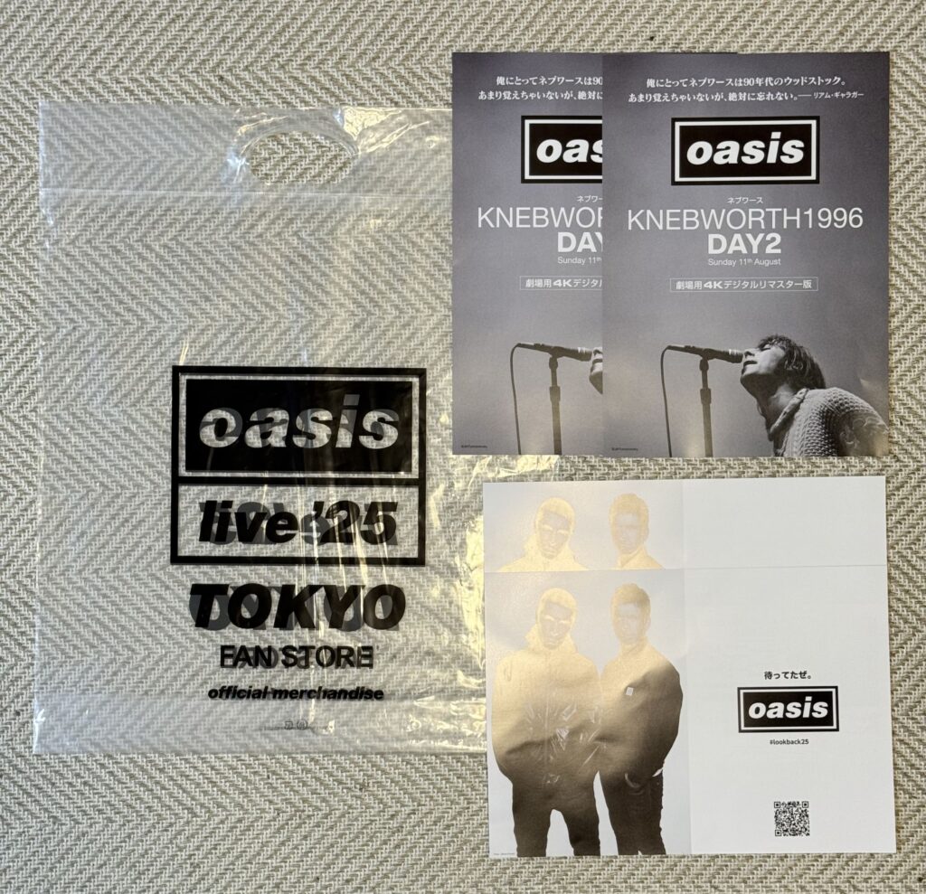 Official merchandise for the British rock band Oasis, including a plastic shopper bag, flyers for the movie 'Oasis: Knebworth 1996', and promotional cards with photos of the band members, all laid out on a beige cloth.