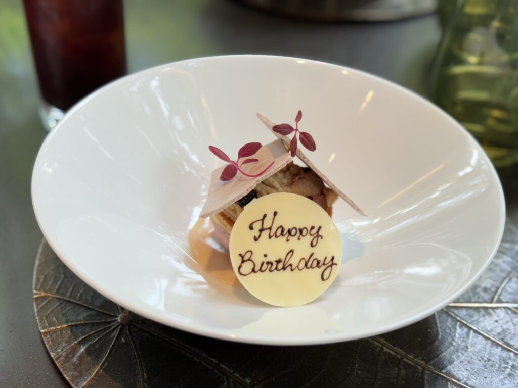 A birthday celebration dessert served on a white plate. It's a Waka-style Mont Blanc, garnished with a white chocolate plaque that reads 'Happy Birthday'.
