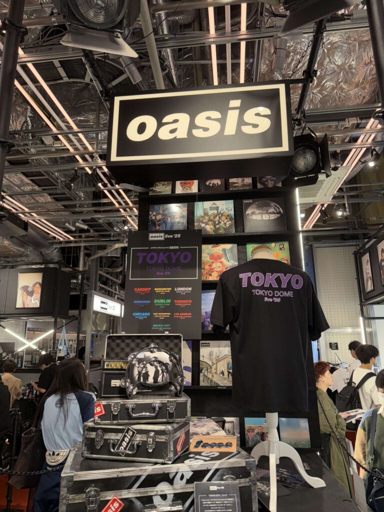 Oasisのポップアップストアの店内の様子。中央にバンドロゴの看板があり、その下には「TOKYO DOME live '25」と書かれたTシャツやレコード、楽器ケースなどが展示されている。