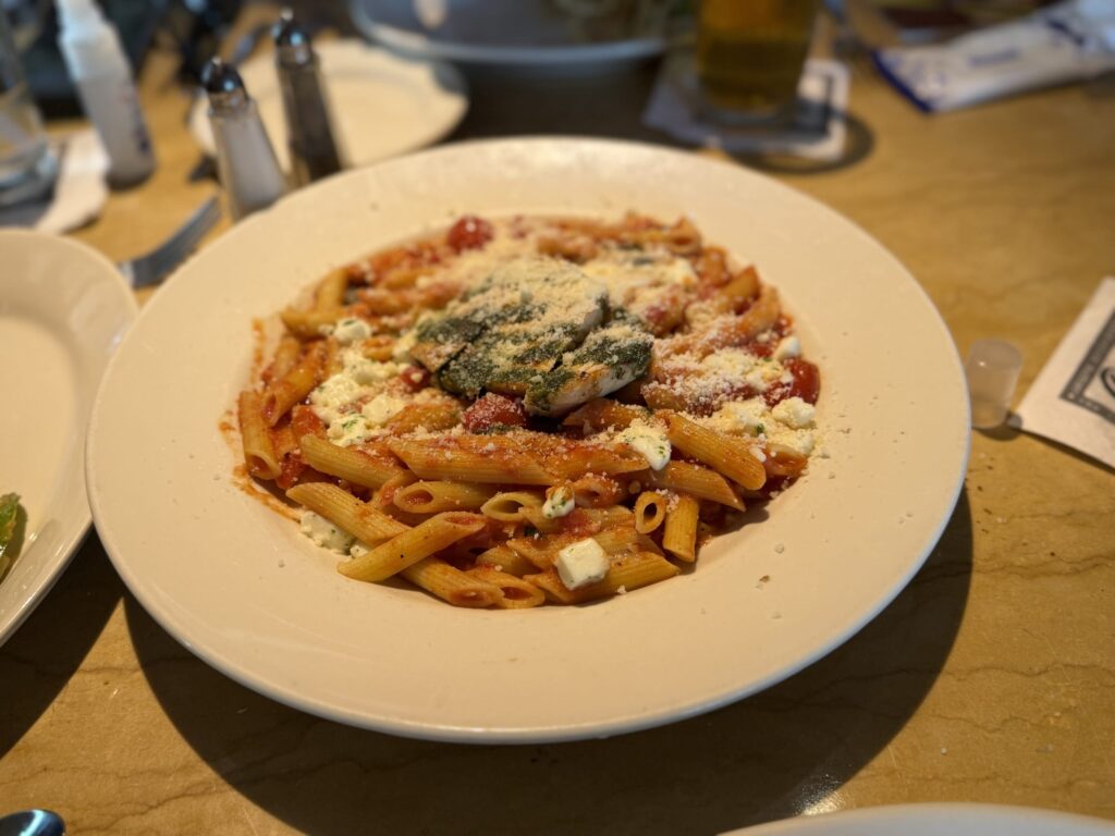A white plate of penne pasta on a restaurant table. It's tossed in a tomato sauce and topped with grated and diced cheese. A slice of chicken with basil pesto is placed in the center.