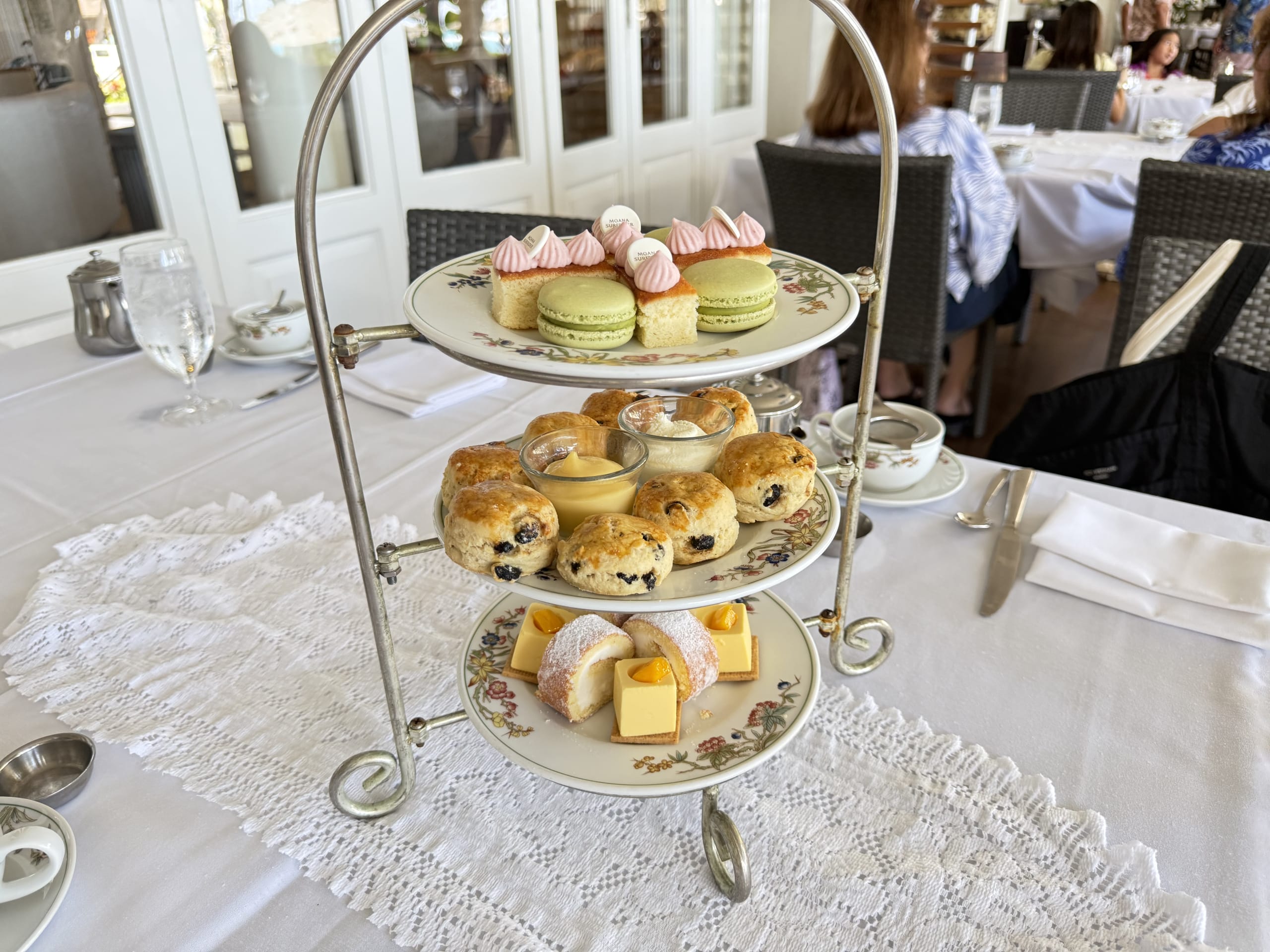 A three-tiered afternoon tea stand on a white tablecloth. The top tier has cakes and macarons, the middle tier has scones, and the bottom tier has a roll cake.