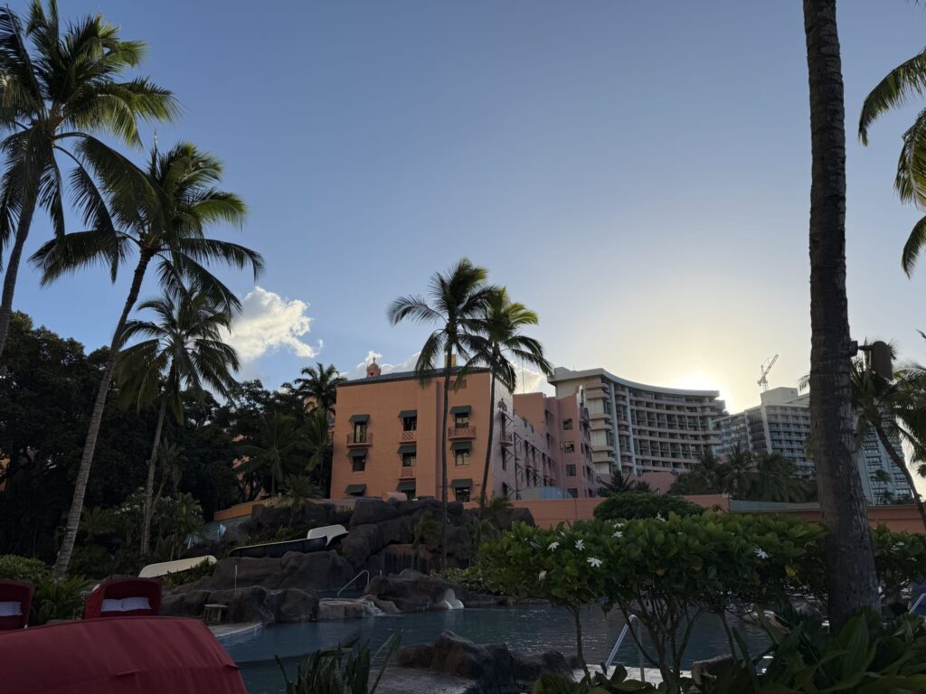 The iconic pink Royal Hawaiian Hotel viewed from the Sheraton Waikiki's pool area, framed by tall palm trees against a clear sky as the sun sets.