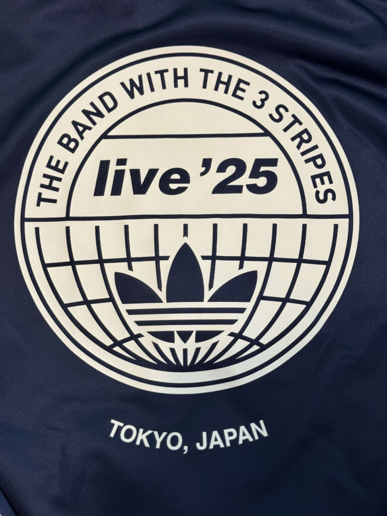 ネイビーの生地に白でプリントされた、アディダスのロゴと「THE BAND WITH THE 3 STRIPES」「live '25」「TOKYO, JAPAN」の文字が入った円形グラフィックのクローズアップ。