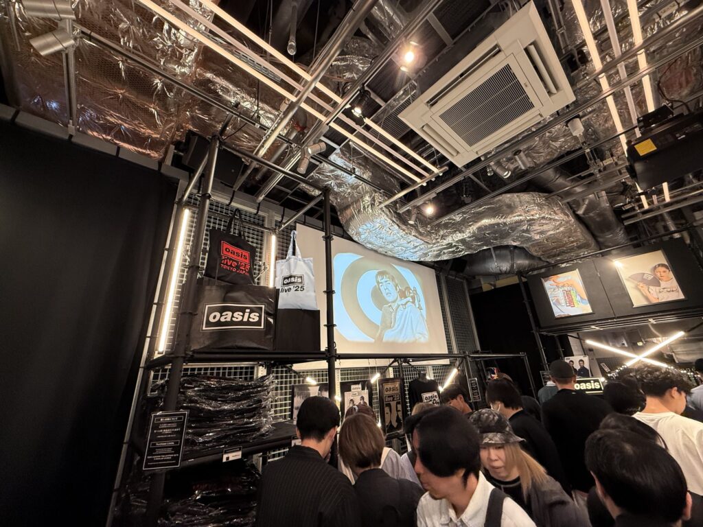 The industrial-style interior of an Oasis pop-up shop. Many fans are eagerly browsing exclusive merchandise like t-shirts and tote bags. A video of a band member is being projected onto a screen in the background.