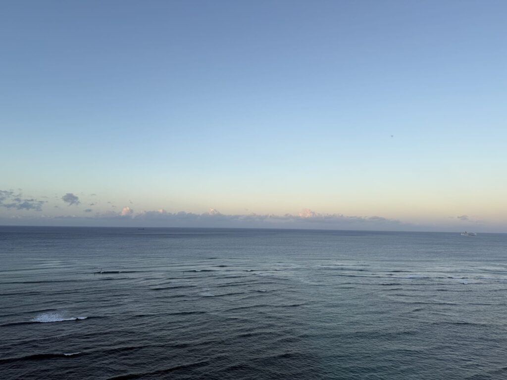 A view from a high floor of the Sheraton showing the calm Waikiki sea under a beautiful morning sky with a soft color gradient. Clouds and distant ships are visible on the horizon.