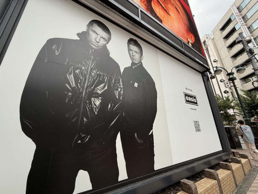 A large black and white billboard on a street in Shibuya, Japan, featuring a photograph of the British rock band Oasis, Liam and Noel Gallagher, posing in dark jackets.