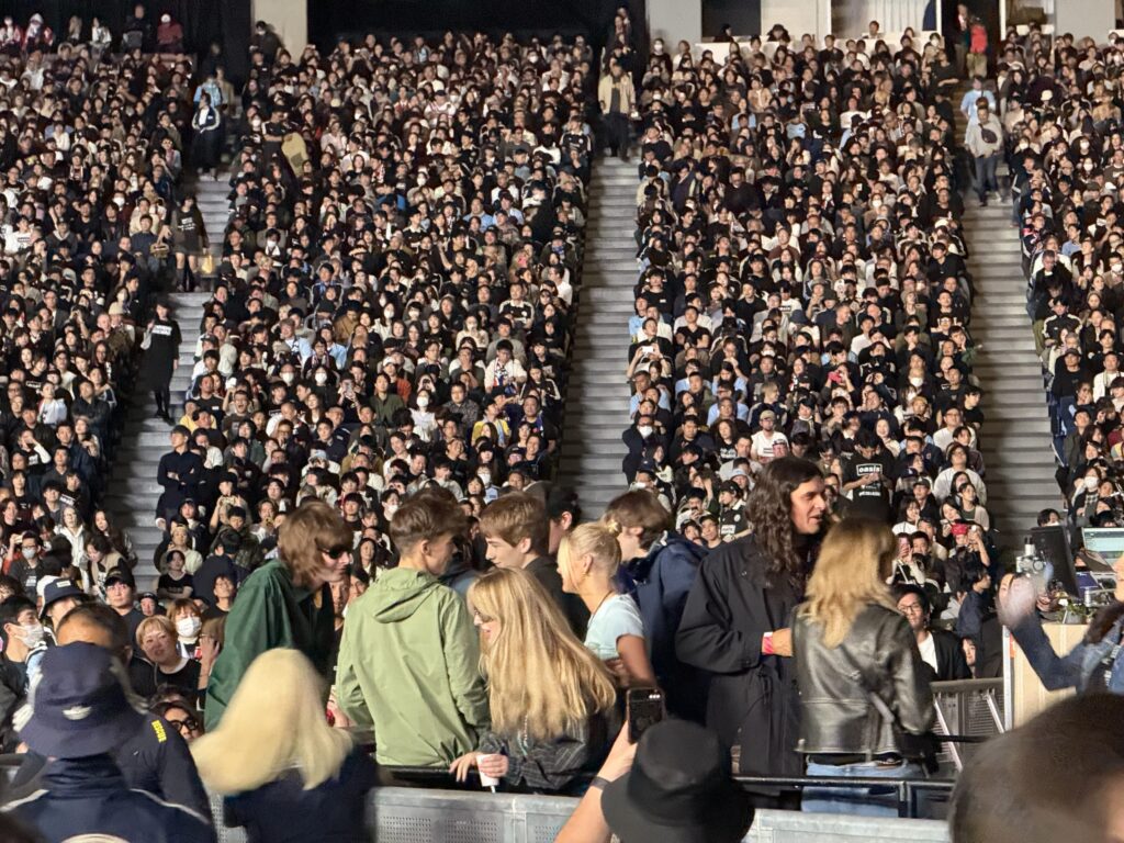 A few people in the special seating area of a concert venue, with a large crowd filling the tiered audience seats behind them.