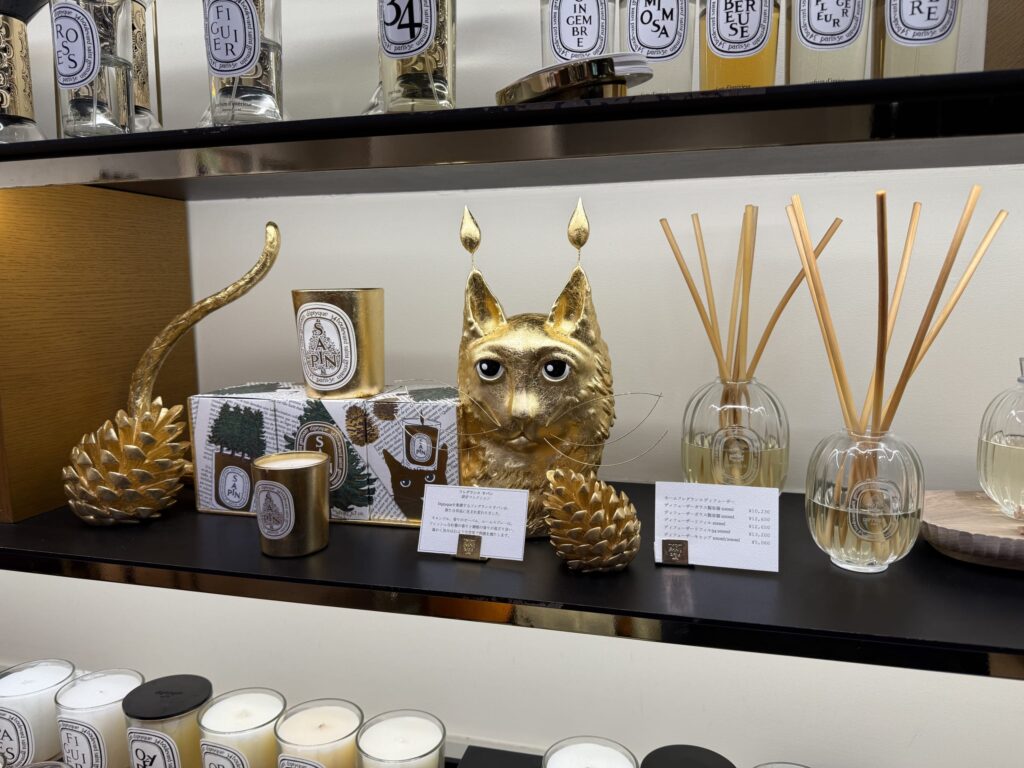 A Diptyque store shelf decorated with a holiday collection. In the center is a golden cat object, surrounded by matching golden candles, pinecones, and a room diffuser.