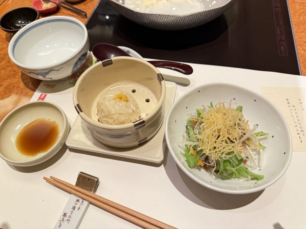 Tofu shumai and a salad placed on a table for a Japanese meal. Chopsticks and a chopstick rest are in the foreground, with small bowls for dipping sauce and serving plates arranged in the background.