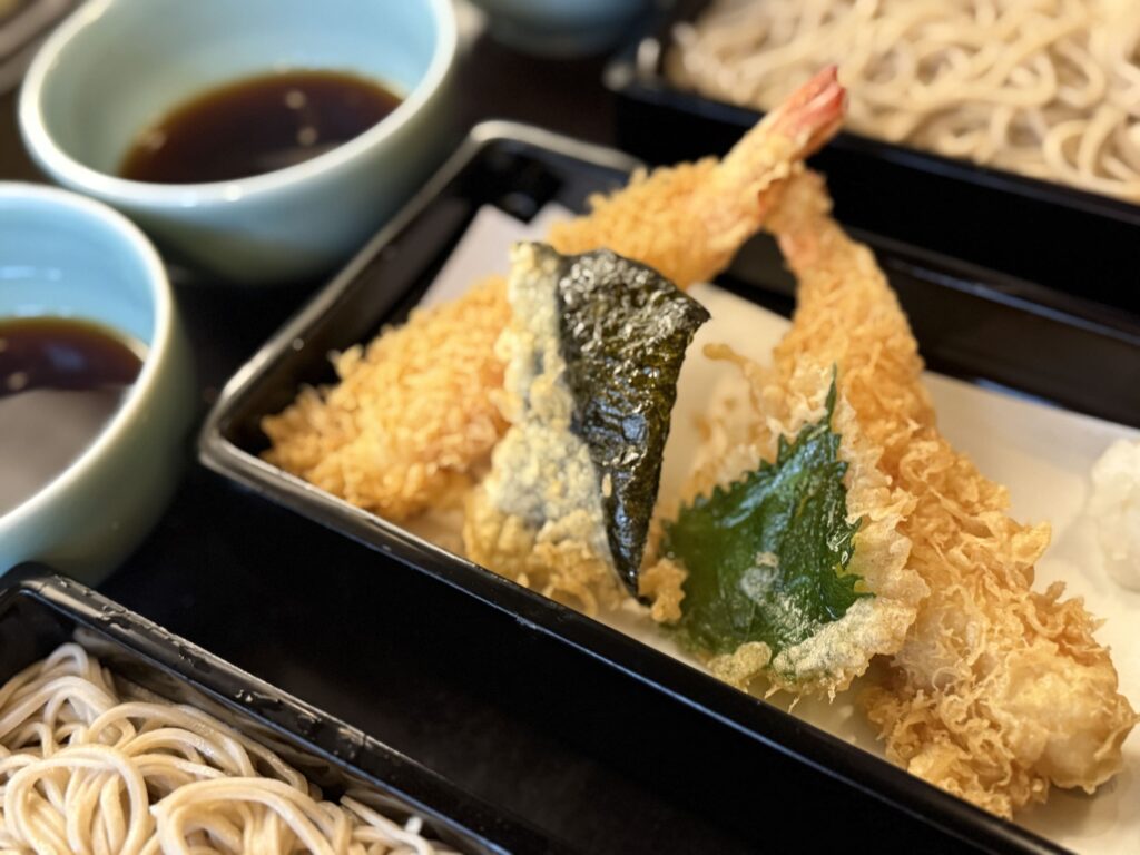 A Japanese meal of Kamo Seiro soba, featuring shrimp tempura, nori tempura, and shiso tempura in a black tray, with cold soba noodles and dipping sauce bowls nearby.