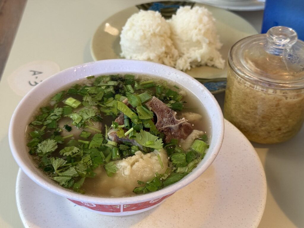 A bowl of oxtail soup generously topped with cilantro, placed on a table. In the background, there is a plate of white rice and a glass jar containing grated ginger.