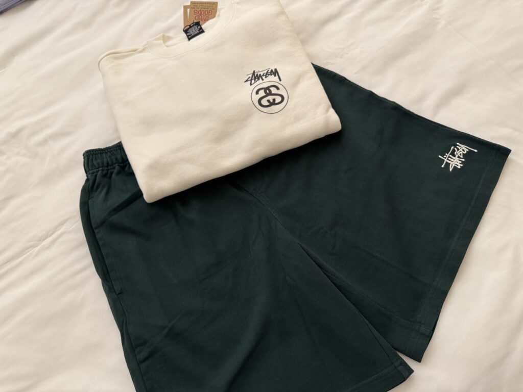 A cream-colored Stüssy sweatshirt and dark green shorts laid out on a white sheet.