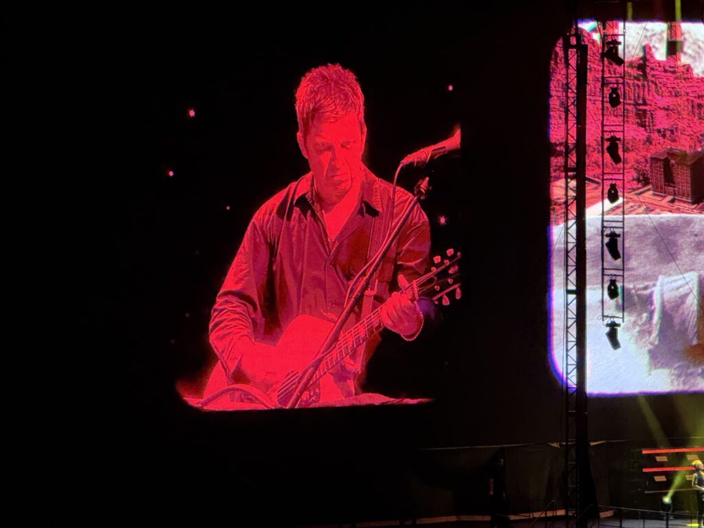 Noel Gallagher playing an acoustic guitar under red lighting, projected onto a giant screen at a concert.