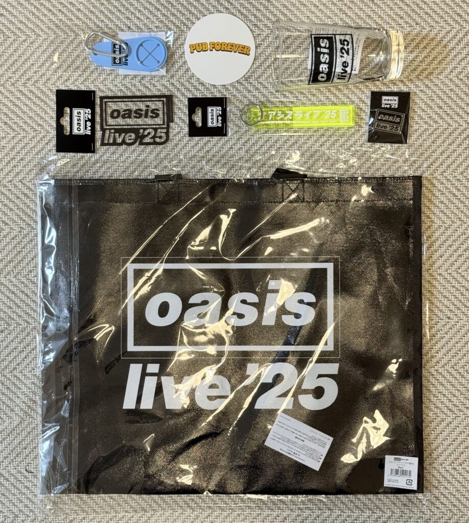 ベージュの布の上に並べられた「オアシス ライブ '25」のグッズ。黒いショッピングバッグ、ビアグラス、ステッカー、ピンバッジ、キーホルダー、PUB FOREVERのコースターなどがある。