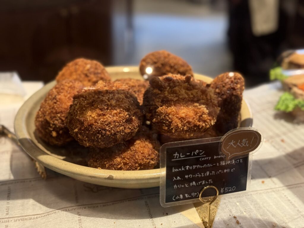A close-up of a ceramic plate piled high with golden-brown, deep-fried Japanese curry bread (kare-pan). A small black sign in front describes the popular item.