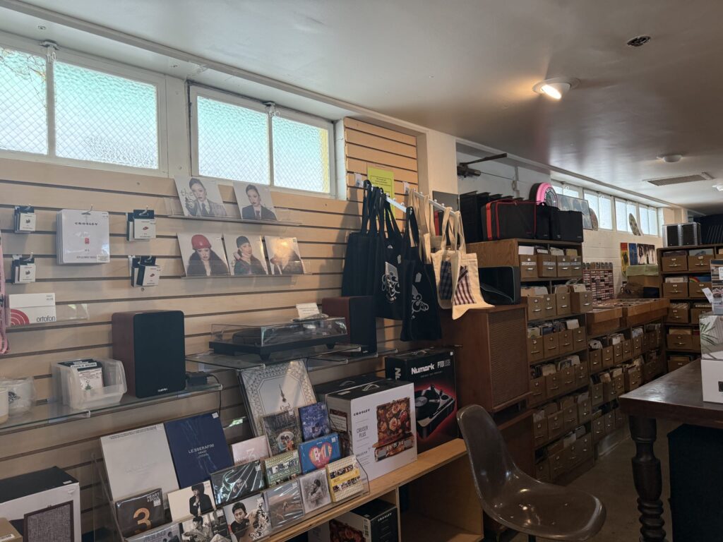 The interior of a record store. Wooden shelves are tightly packed with records, CDs, audio equipment like turntables and speakers, and merchandise such as tote bags.