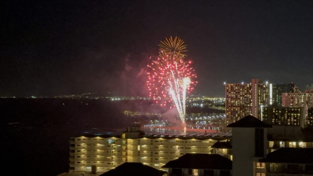 A vibrant red and yellow firework explodes against the night sky over a coastal cityscape.