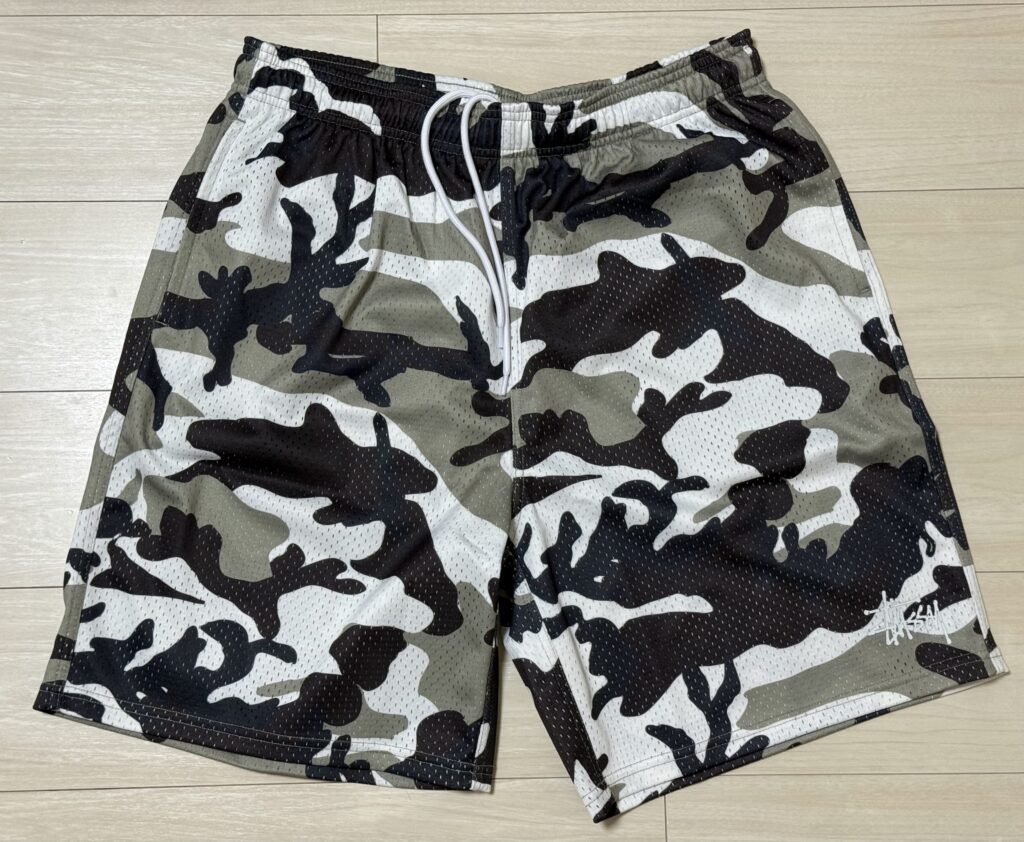 A pair of Stüssy mesh shorts with a white, black, and grey camouflage pattern laid flat on a wood-grain floor.