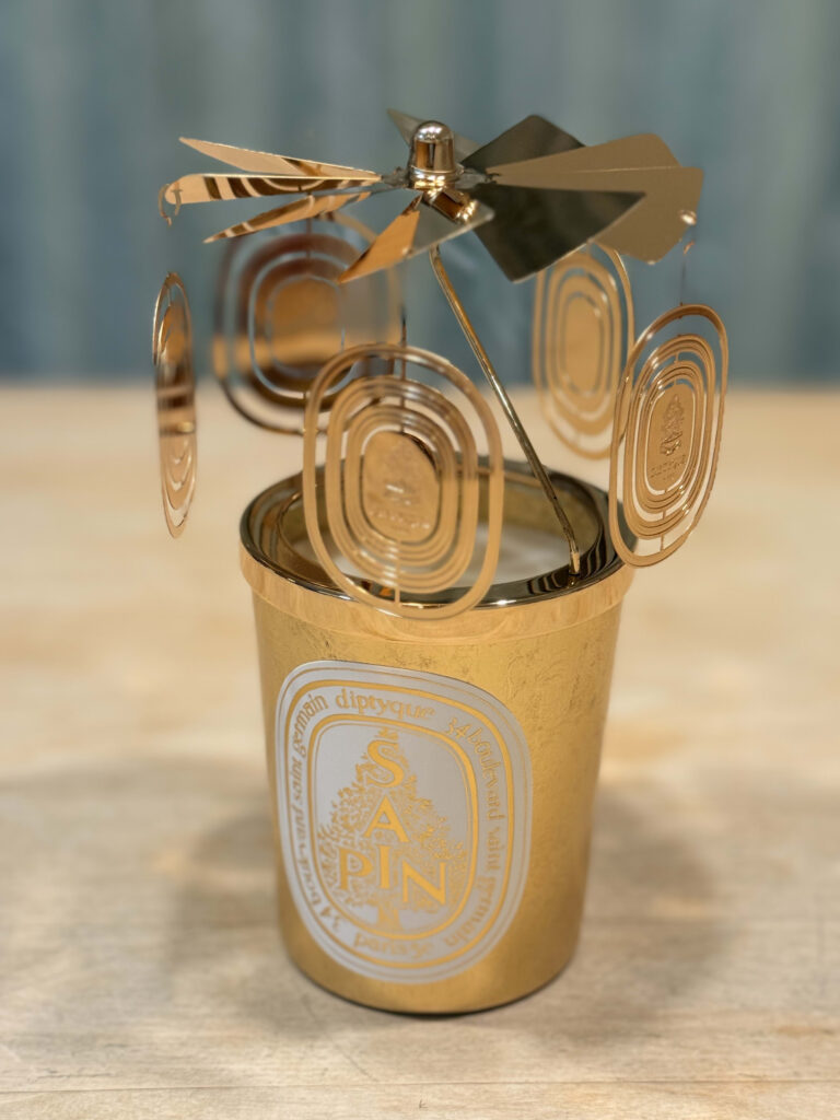 A gold Diptyque 'Sapin' candle with a golden carousel candle topper, placed on a light wooden table.