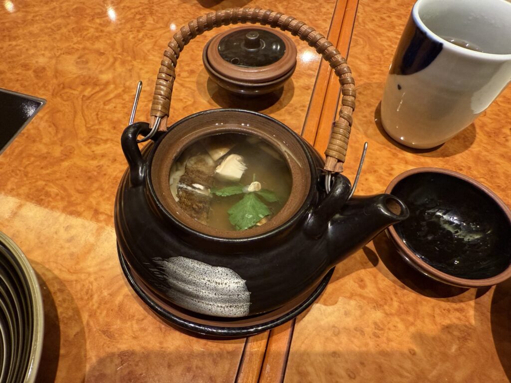 A black ceramic Japanese teapot, a 'dobin', with its lid open, revealing a clear soup with matsutake mushrooms and mitsuba parsley inside, placed on a glossy wood-grain table.