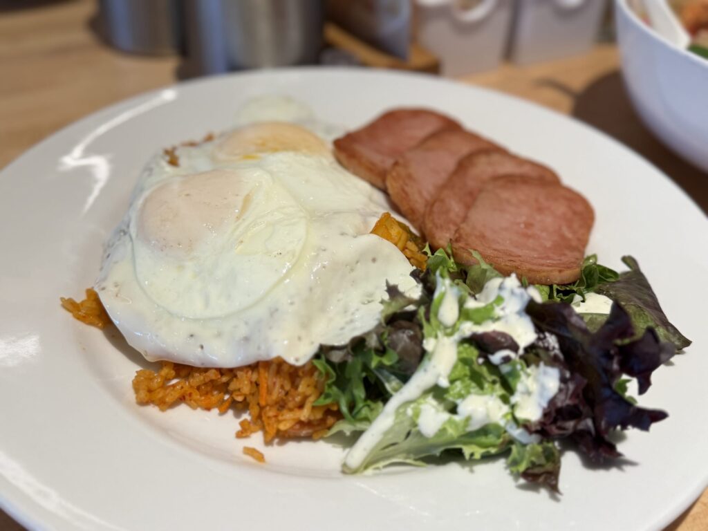 Kimchi fried rice on a white plate, topped with two fried eggs and served with sliced Spam and a green salad.