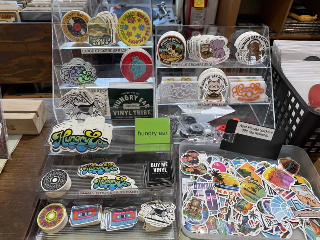 A display stand on the counter of Hungry Ear Records, filled with various original store-branded stickers and an assortment of Hawaii-themed stickers.