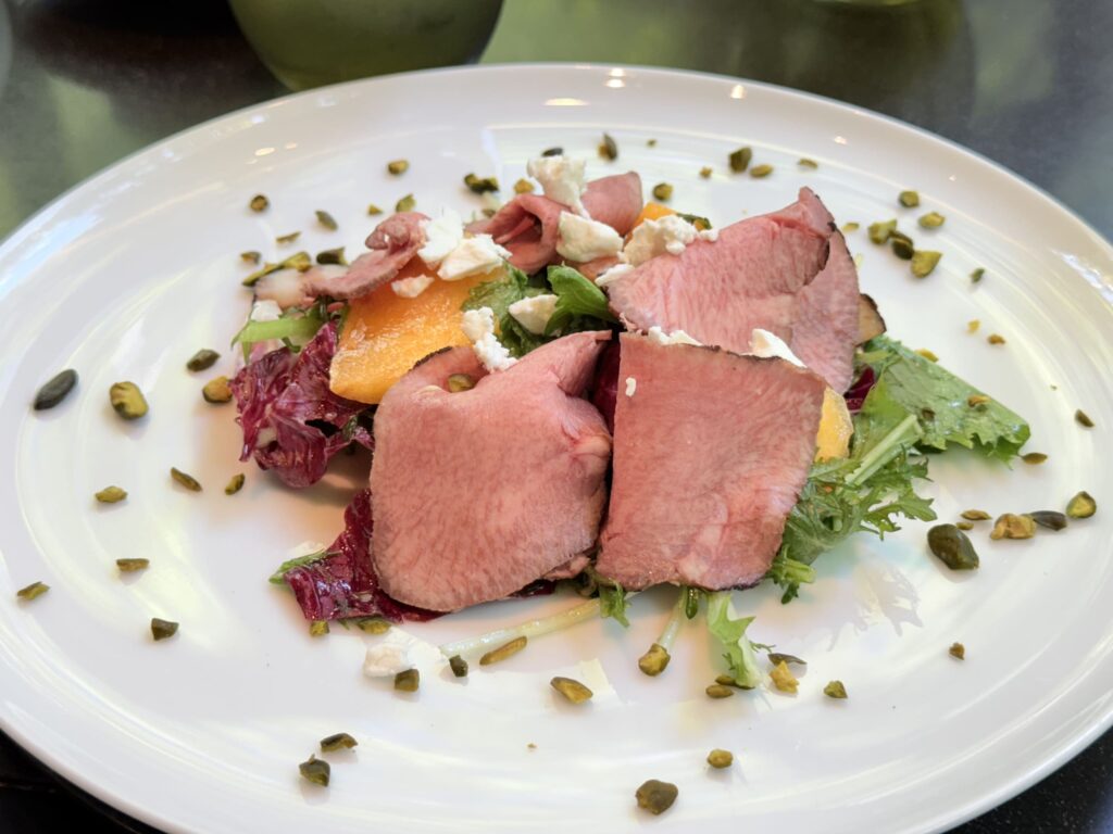 A garden salad with beef tongue confit, persimmon, and Sainte-Maure cheese on a white plate. Chopped pistachios are scattered over the top.