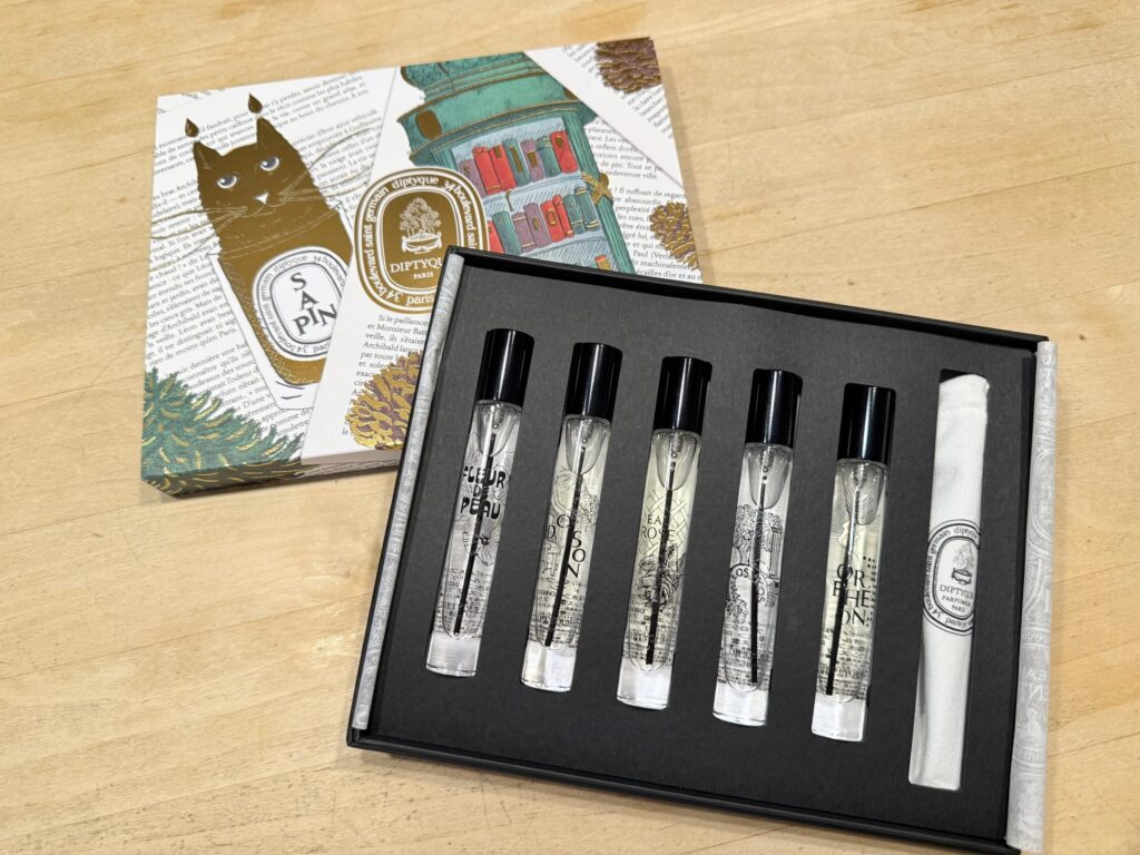 A Diptyque Eau de Parfum discovery set placed on a wooden table. The image shows the decorative box with illustrations of a cat and a bookcase, alongside the open black case containing five travel-sized fragrance sprays.