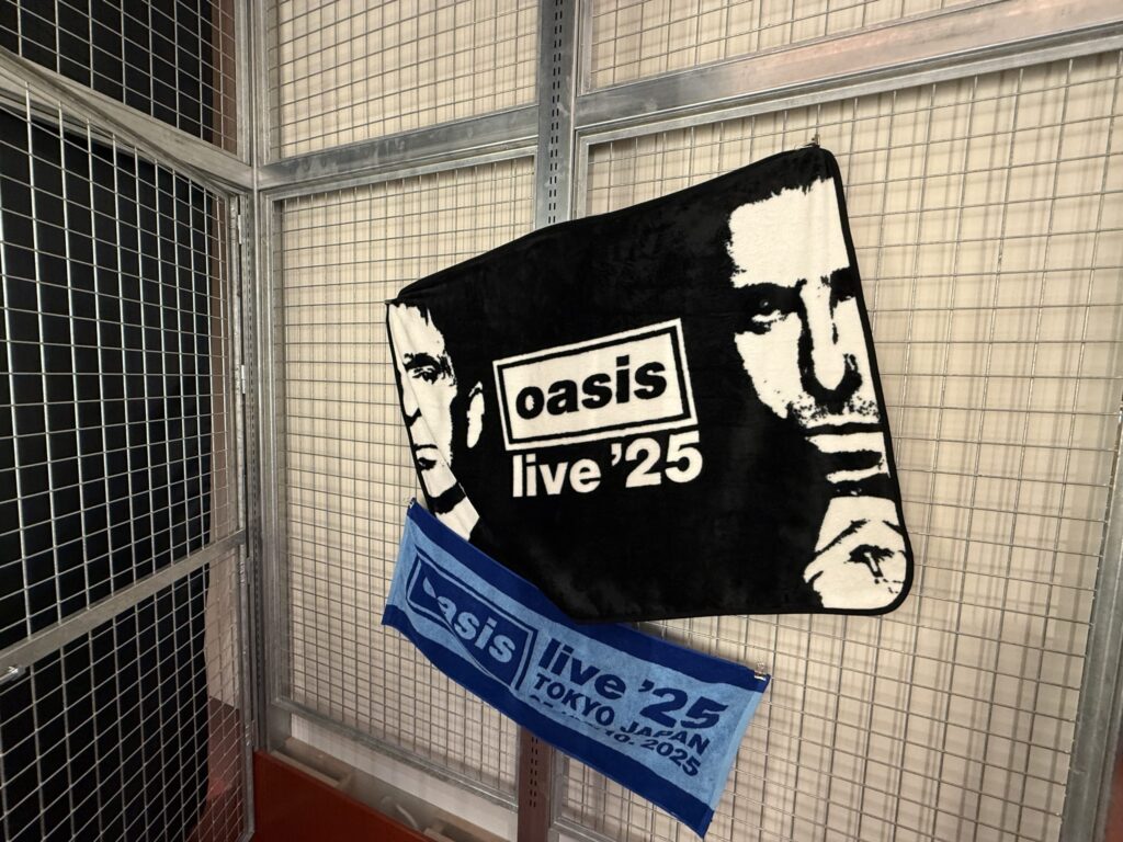 金網の壁に掛けられた、架空のオアシス来日公演「live '25」のグッズタオル。黒いタオルにはリアムとノエルの顔が、青いタオルには「TOKYO JAPAN」の文字がデザインされている。
