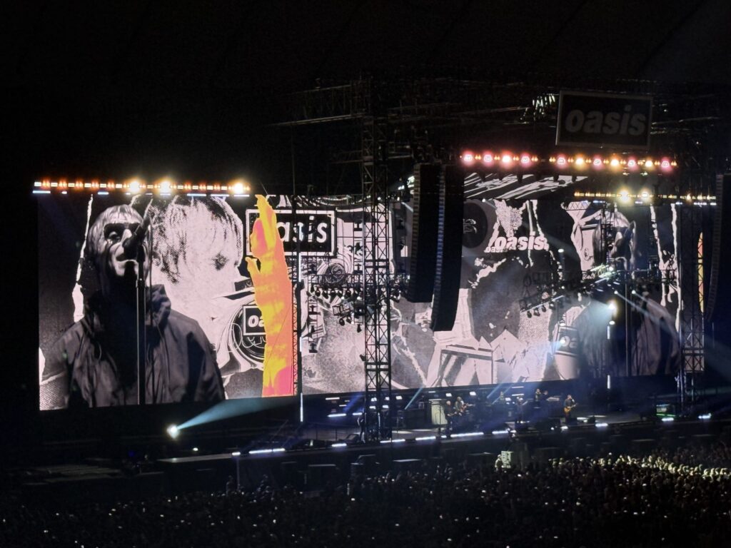 A live concert of the rock band Oasis performing in a stadium. A giant screen in the background displays a black and white photo of vocalist Liam Gallagher, with a massive crowd of spectators below the stage.