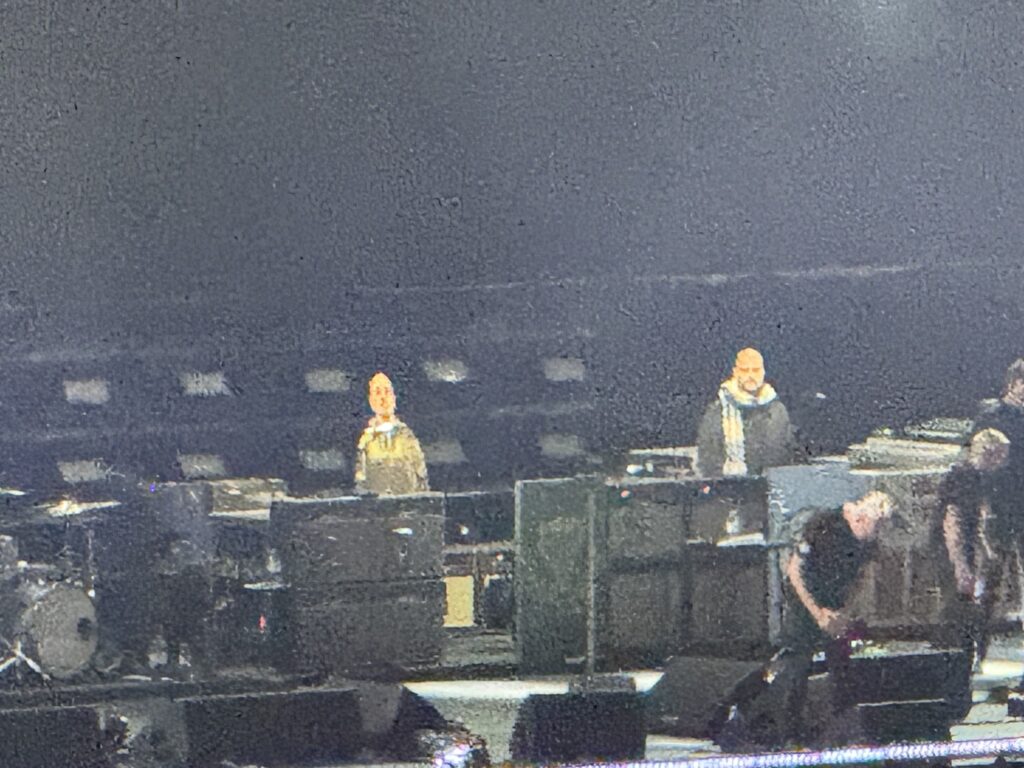 On a dark concert stage, former Oasis guitarist Bonehead and another similar-looking bald man — Manchester City manager Betts — stand side by side among Liam Gallagher's band members.