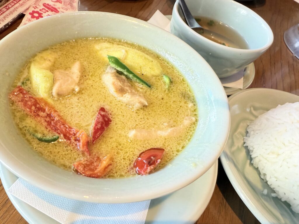 A bowl of Thai green curry with chicken and vegetables, served with a side of white rice and a small bowl of soup on a wooden table.
