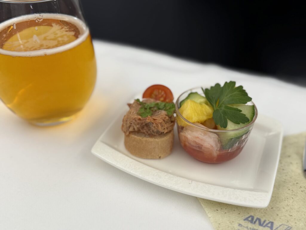 白いテーブルクロスの上に置かれた、ANAの機内食。グラスに注がれたビールと、豚肉のリエットのカナッペ、シーフードとフルーツのサラダが白いお皿に盛り付けられている。