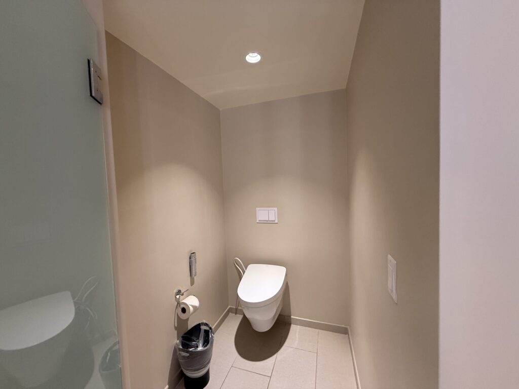 A clean and spacious hotel toilet room with beige walls, featuring a wall-mounted toilet with a bidet seat.