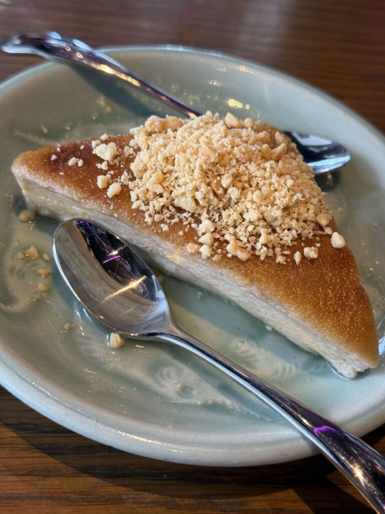 A triangular slice of taro pudding cake topped with crushed nuts, served on a light green plate with two spoons.
