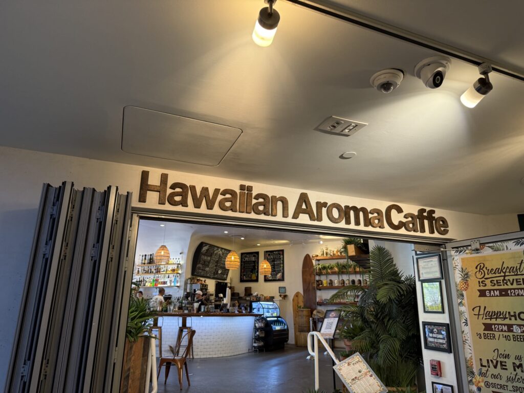 「Hawaiian Aroma Caffe」と木製の文字で書かれた看板があるカフェの入り口。ドアは開け放たれ、カウンターや観葉植物がある店内の様子が見える。
