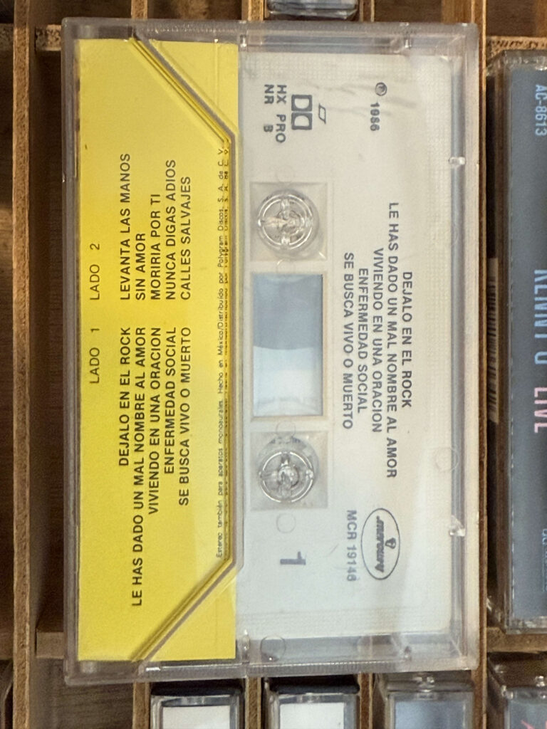 A close-up of a vintage cassette tape in its clear case, resting in a wooden storage rack. The J-card is bright yellow with a tracklist in Spanish.