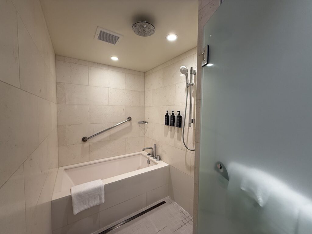 A hotel bathroom covered in light beige tiles. There is a bathtub in the back, with a frosted glass door and shower area visible in the foreground. A rain shower head is installed on the ceiling.