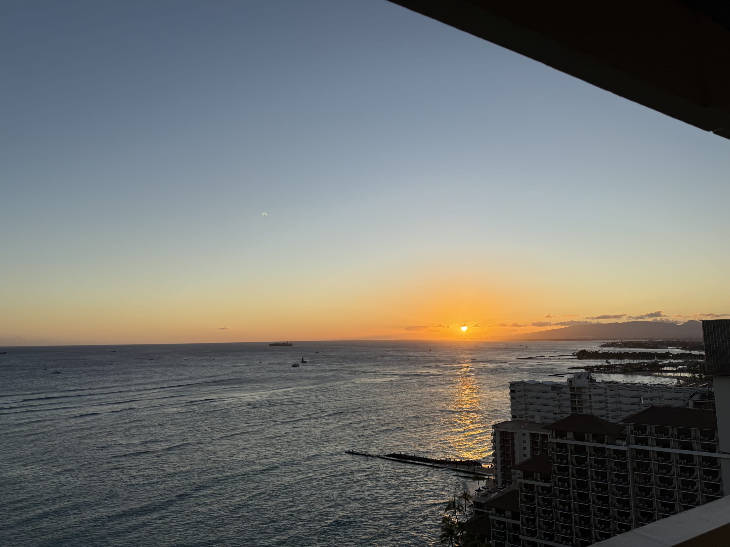 Sunset over the ocean viewed from the lanai of the Sheraton Waikiki hotel. The orange sun illuminates the sea, with the coastline and mountains visible in the distance.
