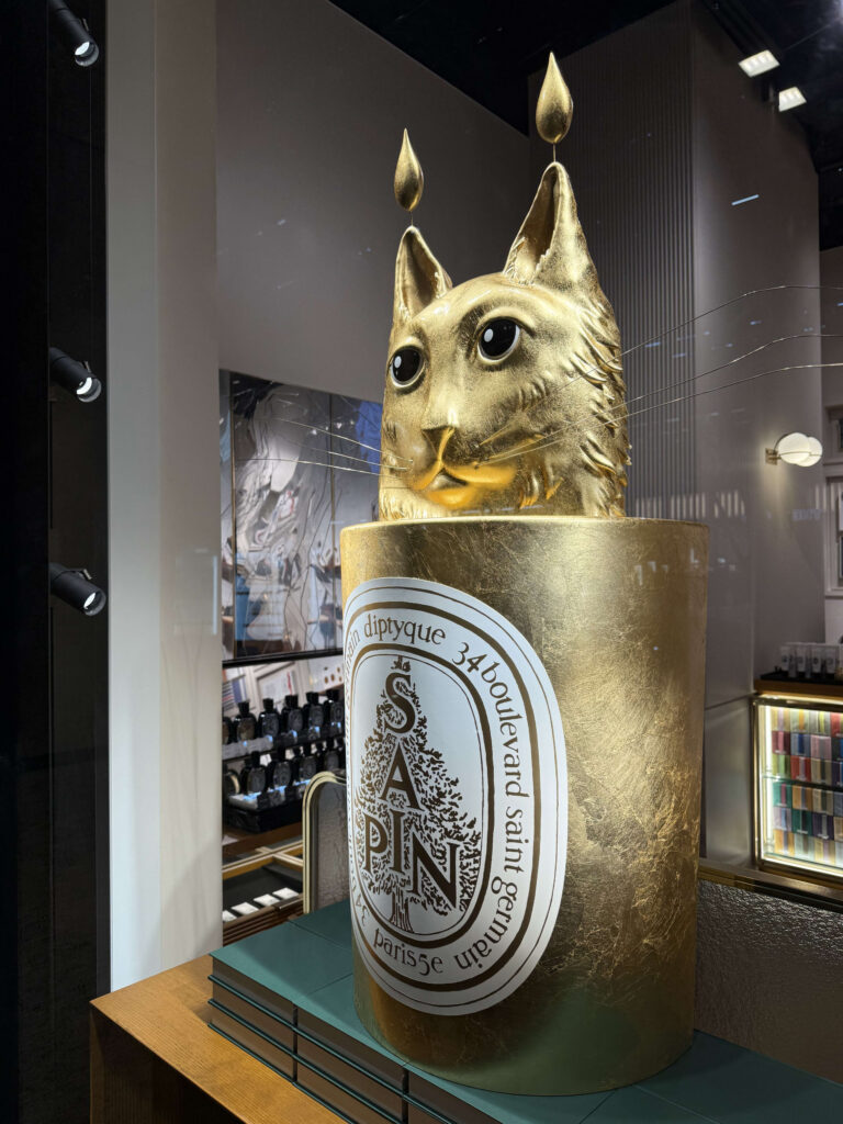 A store display featuring a large, golden sculpture of a cat's head emerging from an oversized Diptyque candle with a 'Sapin' label.