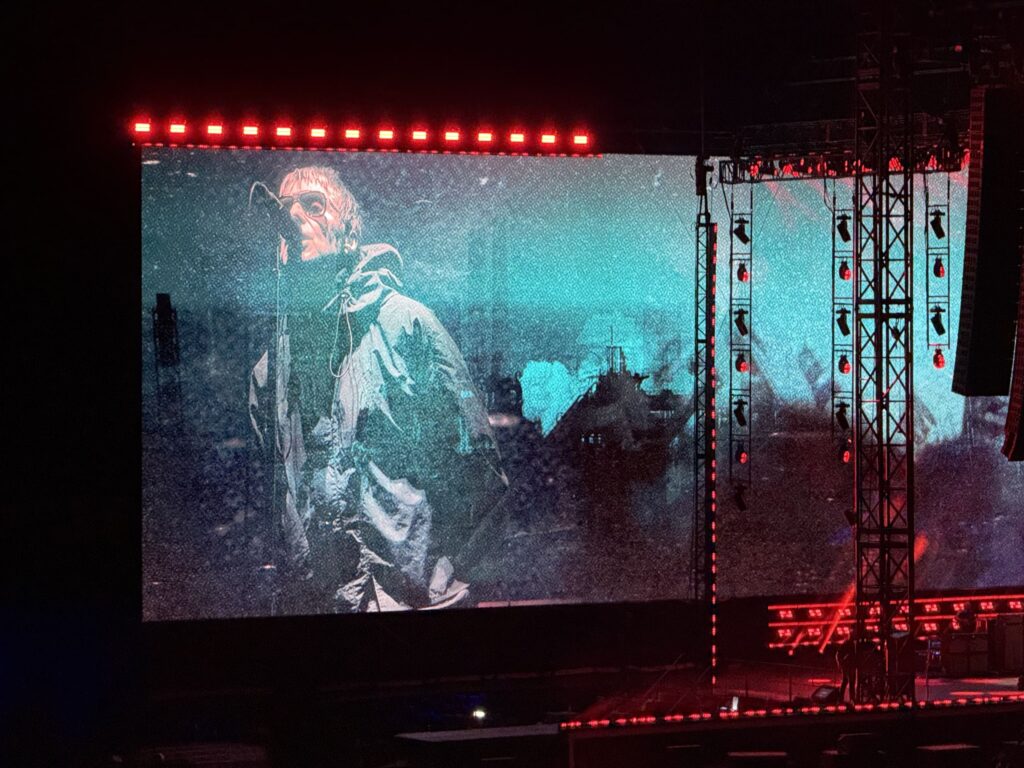 A large screen at a concert shows a man in a parka and sunglasses singing into a microphone. Stage rigging and red lights frame the screen.