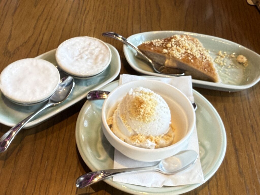 Three kinds of desserts served on light green tableware on a wooden table: a slice of taro pudding cake, a scoop of coconut ice cream, and two cups of coconut pudding.