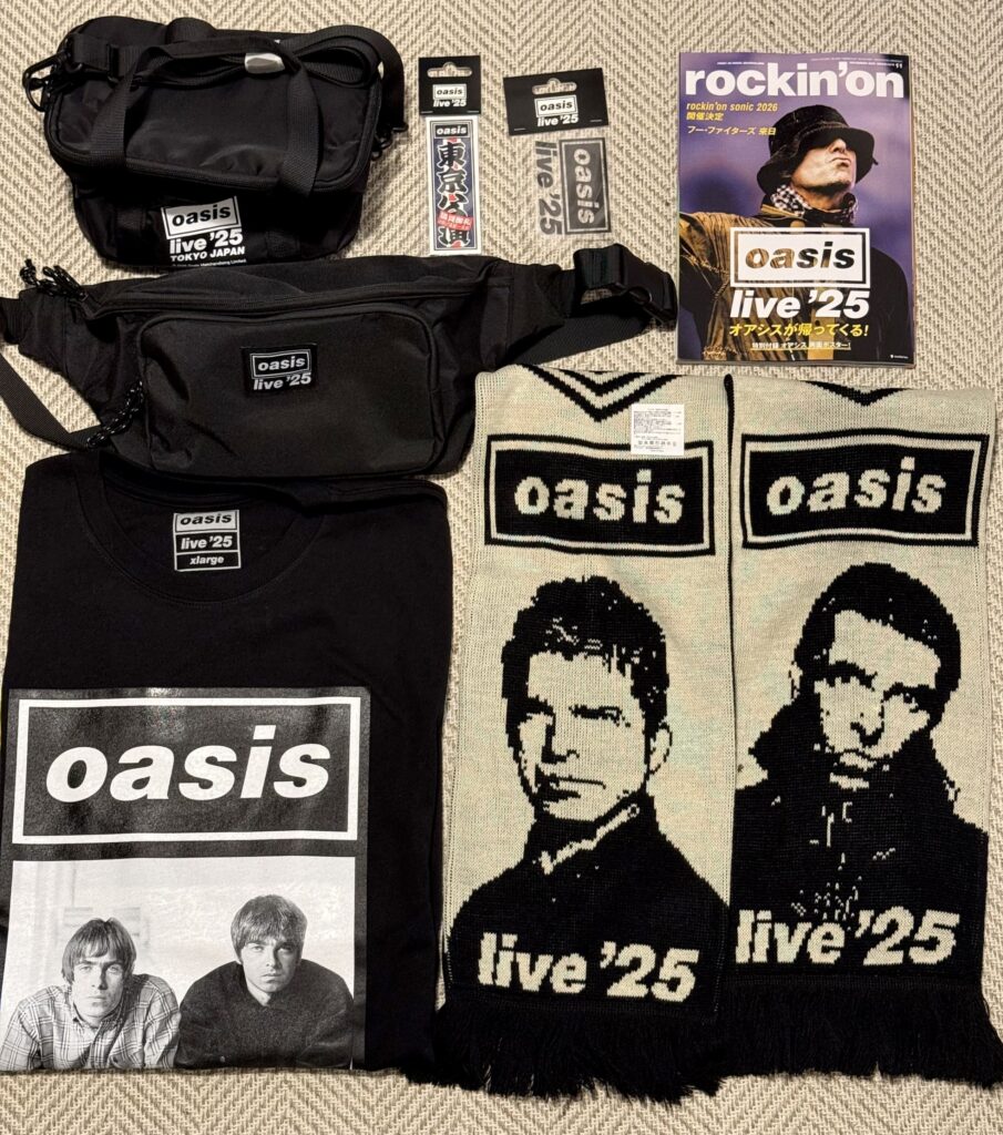 Oasisの「live '25」ツアーグッズ。Tシャツ、ショルダーバッグ、ウエストポーチ、ニットマフラー、ステッカー、そして雑誌が床に並べられている。