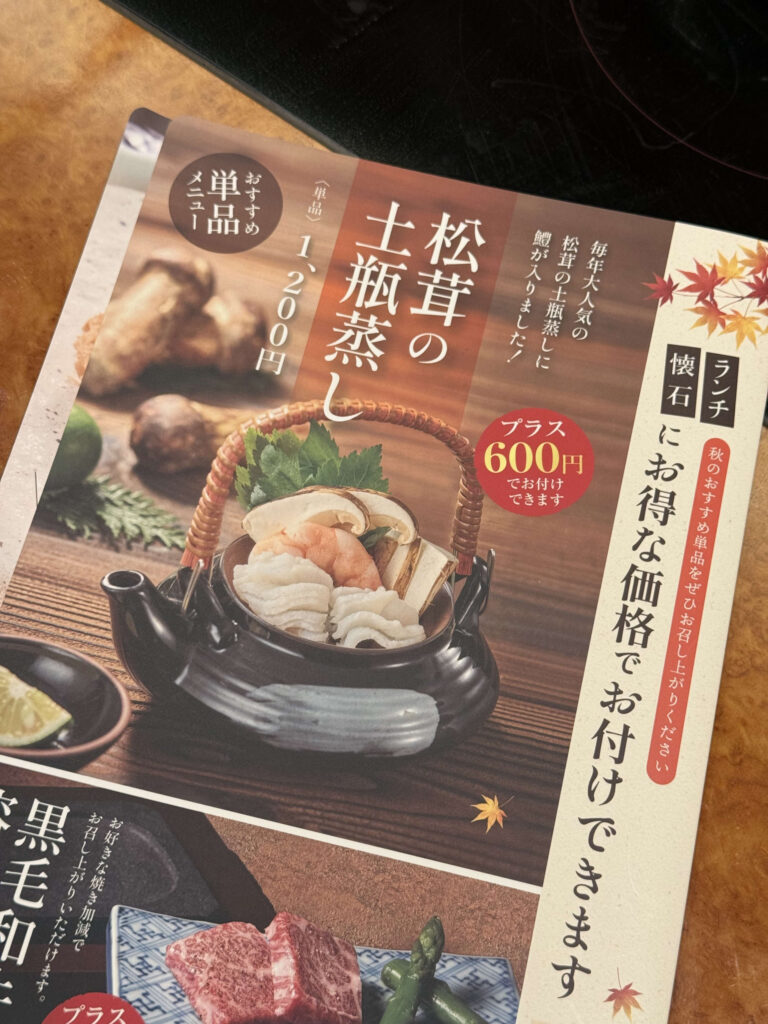木製のテーブルに置かれた、松茸の土瓶蒸しを紹介するレストランの秋メニュー。黒い土瓶に入った料理の写真が大きく写っている。