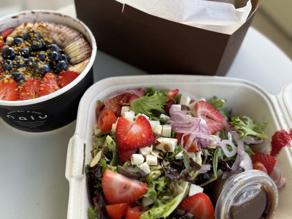 A takeout lunch on a white table, featuring a salad with strawberries and cheese, and an acai bowl topped with fruit.
