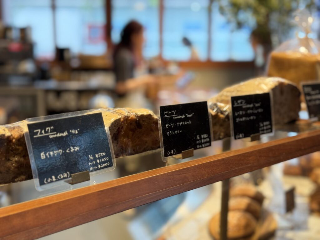 A close-up of sourdough loaves on display at a bakery. The focus is on the bread in the foreground, with handwritten labels in Japanese for