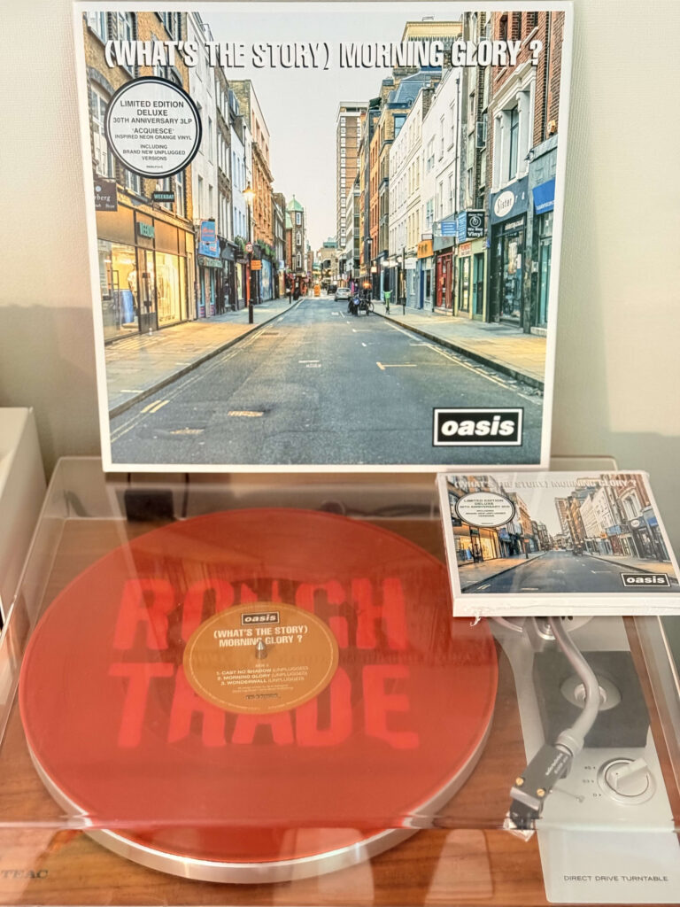 ターンテーブルで再生されているオアシスのアルバム「(What's the Story) Morning Glory?」30周年記念限定盤のネオンオレンジのレコード。背景にはアルバムジャケットとCDが飾られている。