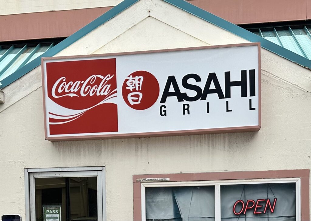 The sign for the Asahi Grill restaurant on an exterior wall. It features the Coca-Cola logo, the Japanese kanji for 'asa' in a red circle, and the name 'ASAHI GRILL' in black letters.